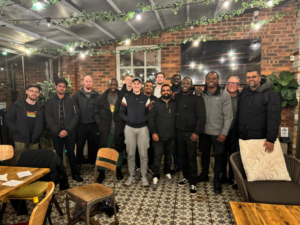 A-group-of-diverse-men-standing-together-in-a-cozy-indoor-setting-with-greenery-and-string-lights-overhead-smiling-and-posing-for-a-group-photo-representing-a-close-knit-community-gathering