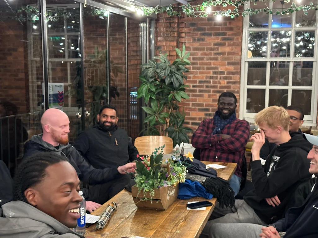 group-of-men-sitting-around-a-table-laughing-and-smiling-in-a-relaxed-setting-with-brick-walls-and-plants-in-the-background