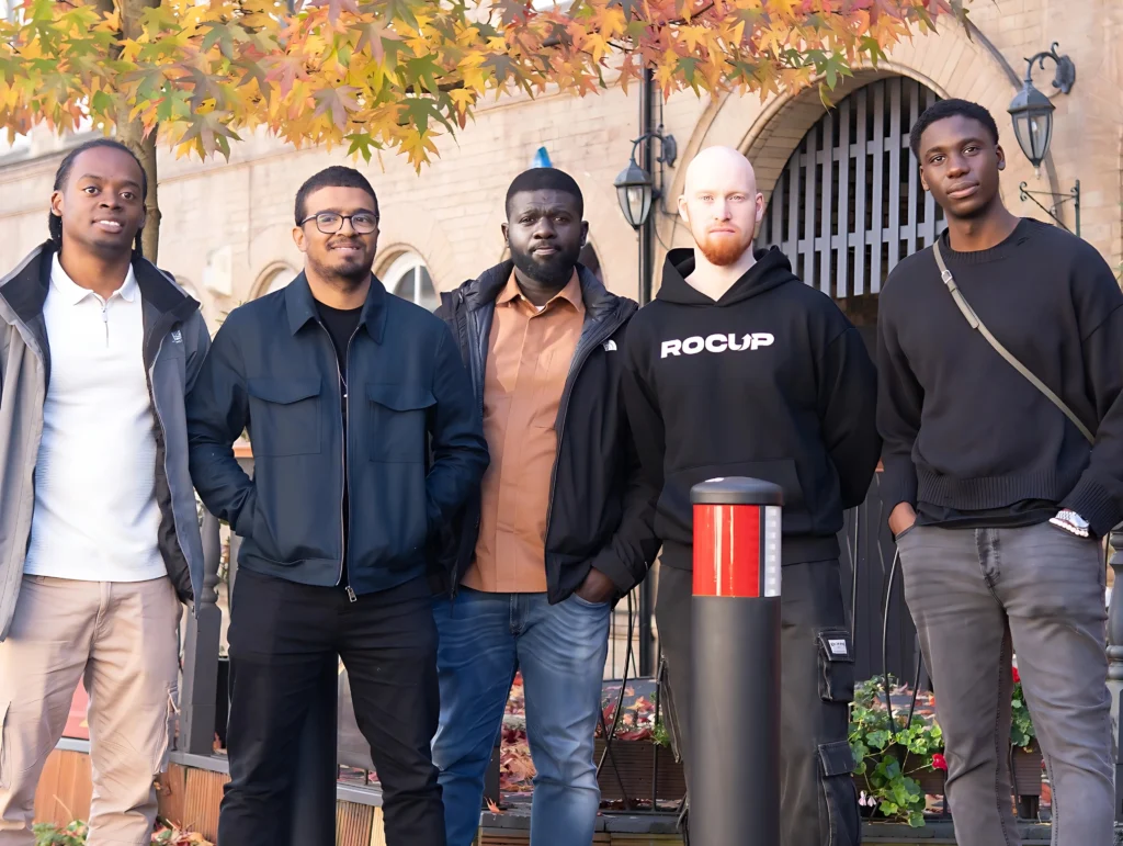 A-group-of-five-men-standing-outdoors-in-front-of-a-brick-building-with-autumn-leaves-in-the-background