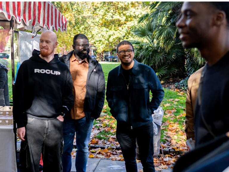 group-of-men-standing-outdoors-smiling-near-market-stall-surrounded-by-autumn-leaves-and-trees