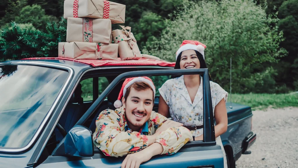 A-man-and-woman-wearing-Santa-hats-smiling-with-a-car-loaded-with-Christmas-presents-and-a-tree