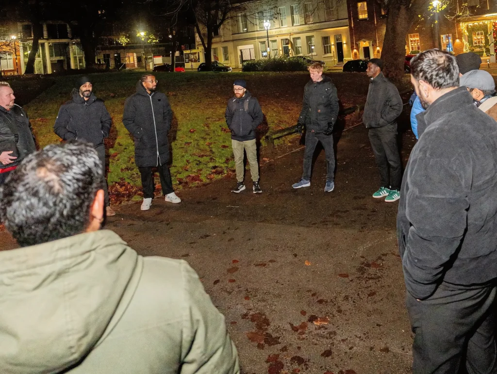 MPC-group-standing-in-a-circle-outdoors-at-night-wearing-warm-clothing-with-a-dimly-lit-park-and-buildings-in-the-background