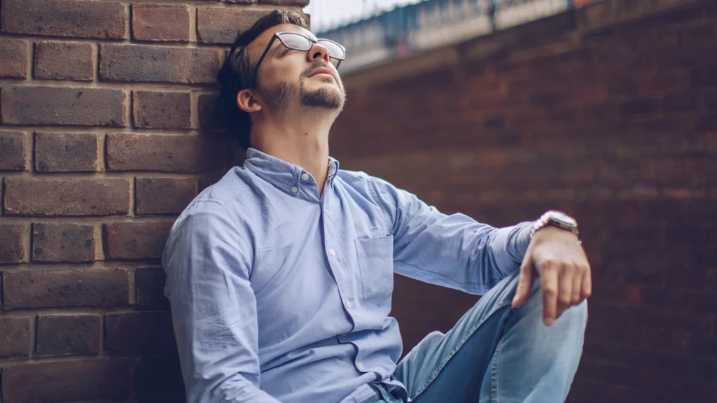 A-man-sitting-against-a-brick-wall-with-his-head-tilted-back-and-eyes-closed-symbolizing-social-anxiety-and-reflection