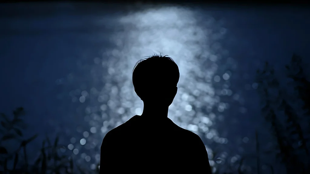 A-silhouette-of-a-man-staring-at-a-reflective-body-of-water-under-moonlight-symbolising-the-silent-struggle-of-inner-thoughts-and-emotions