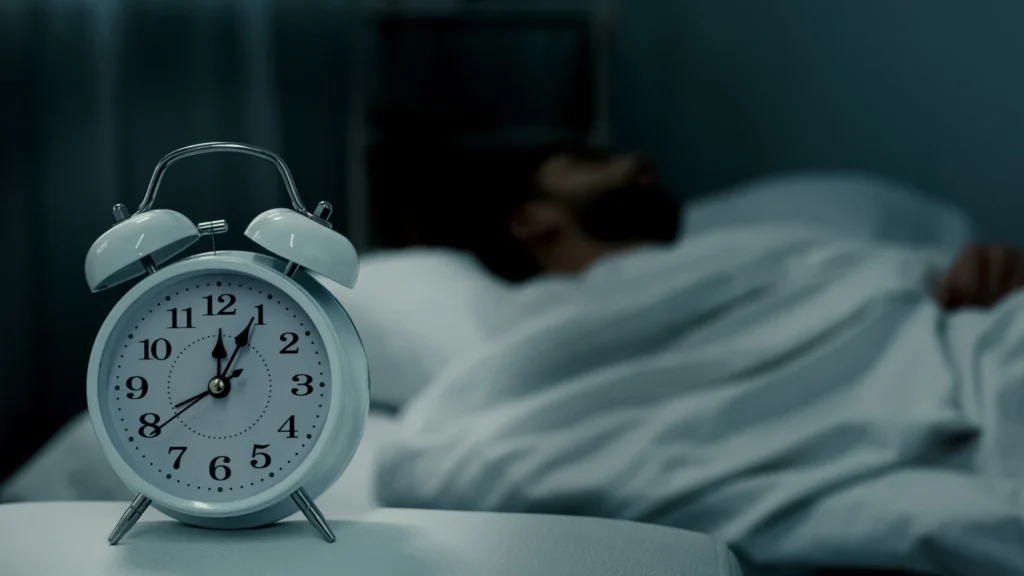 A-person-sleeping-in-bed-with-an-alarm-clock-showing-past-12-AM-visually-raising-the-question-Is-6-Hours-of-Sleep-Enough