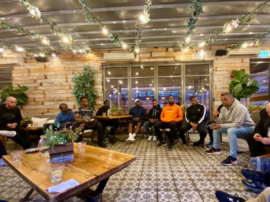 A-group-of-men-seated-in-a-circle-during-a-Mens-Prosperity-Club-MPC-event-engaged-in-a-conversation-in-a-warmly-lit-indoor-venue-with-hanging-lights-greenery-and-tiled-flooring