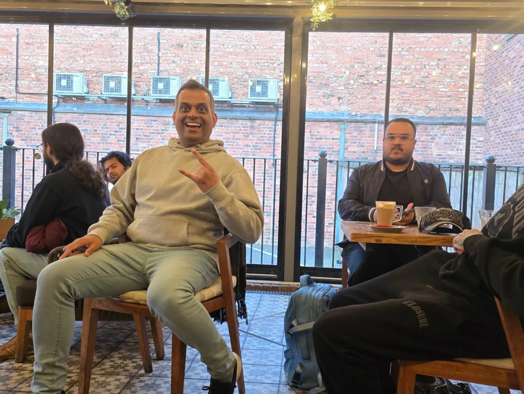 A-man-sitting-at-a-table-in-a-relaxed-café-setting,-giving-a-thumbs-up-while-another-man-sits-quietly-with-coffee