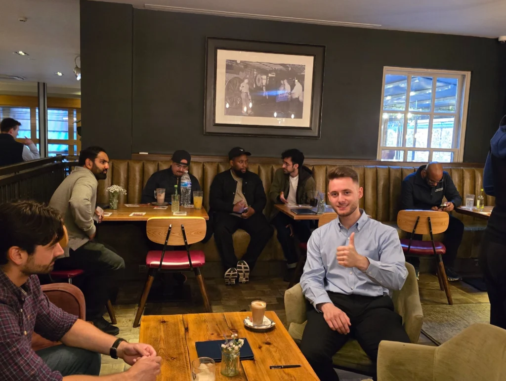 Group-of-men-seated-in-a-cafe,-with-one-man-in-the-foreground-giving-a-thumbs-up-and-a-friendly-smile