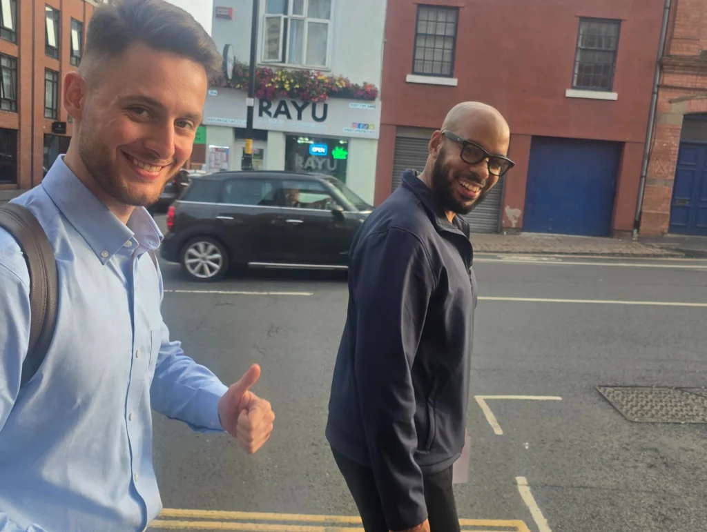 Image-of-two-men-walking-on-a-street,-smiling-and-giving-a-thumbs-up,-with-a-RAYU-sign-in-the-background