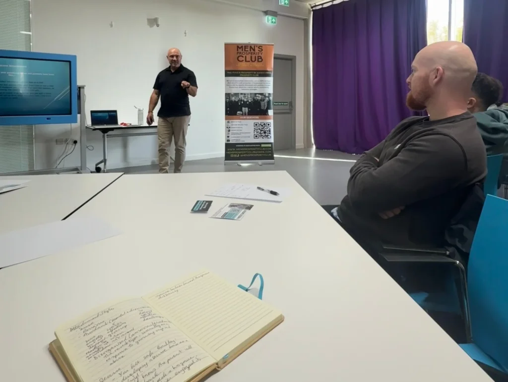 A-view-of-the-PD-workshop-where-John-Kenny-is-presenting-and-an-audience-member-is-taking-notes-with-the-Men's-Prosperity-Club-banner-in-the-background